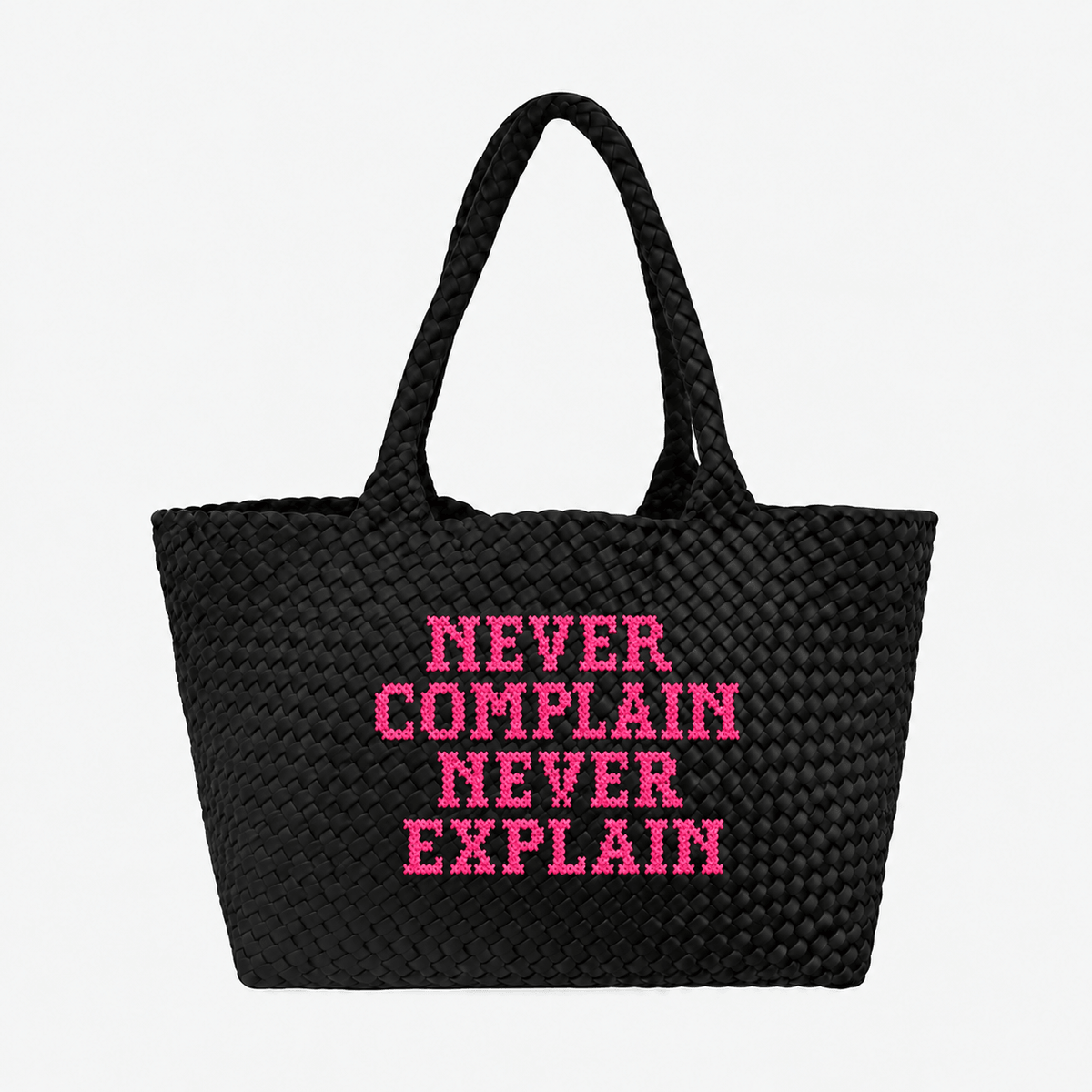 A black woven straw tote bag with two handles, featuring the text 'NEVER COMPLAIN NEVER EXPLAIN' printed in bright pink capital letters on the front, set against a plain white background.